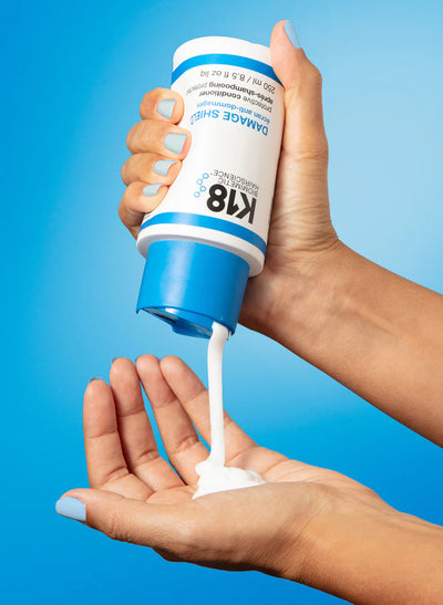 Hand holding DAMAGE SHIELD Conditioner bottle, dispensing creamy product onto palm against blue background.