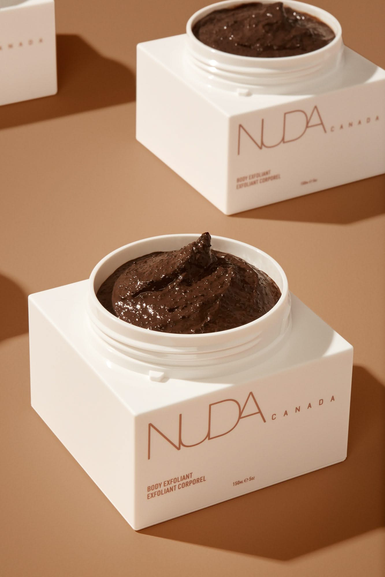 Nuda Canada Body Exfoliant in a white jar with brown exfoliating product, ideal for prepping skin for sunless tanning.