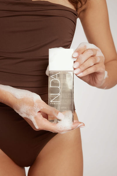 Woman holding Nuda body exfoliant bottle with foam, showcasing skin prep for sunless tanning.