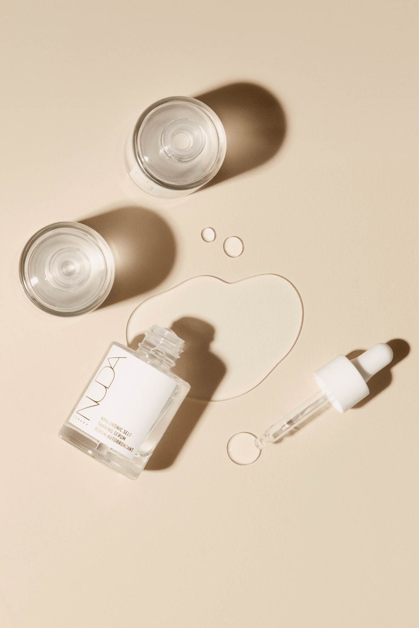 Clear skincare serum bottle with dropper, two glass jars, and liquid drops on a beige background.