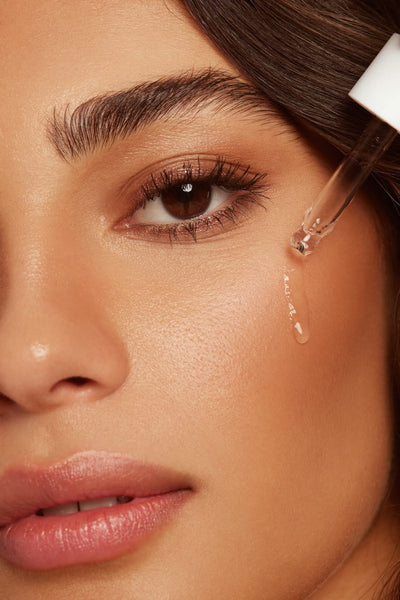 Close-up of a woman's face applying a facial self-tanner drop for a luminous glow.