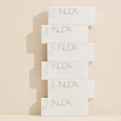 Stacked self-tanning lotion bottles featuring NUDA branding on a neutral background.