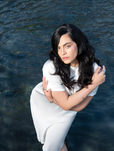 Woman in a white dress poses gracefully by water, showcasing her long, wavy hair and serene expression.