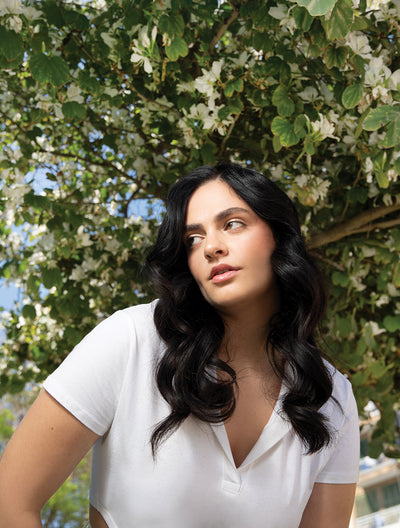 Woman with long, wavy black hair wearing a white shirt, posing in front of a blooming tree.