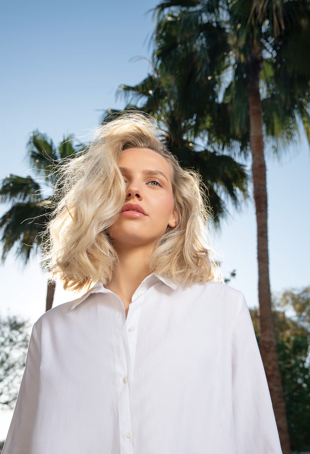 Young woman with wavy blonde hair wearing a white shirt, standing under palm trees against a sunny sky.