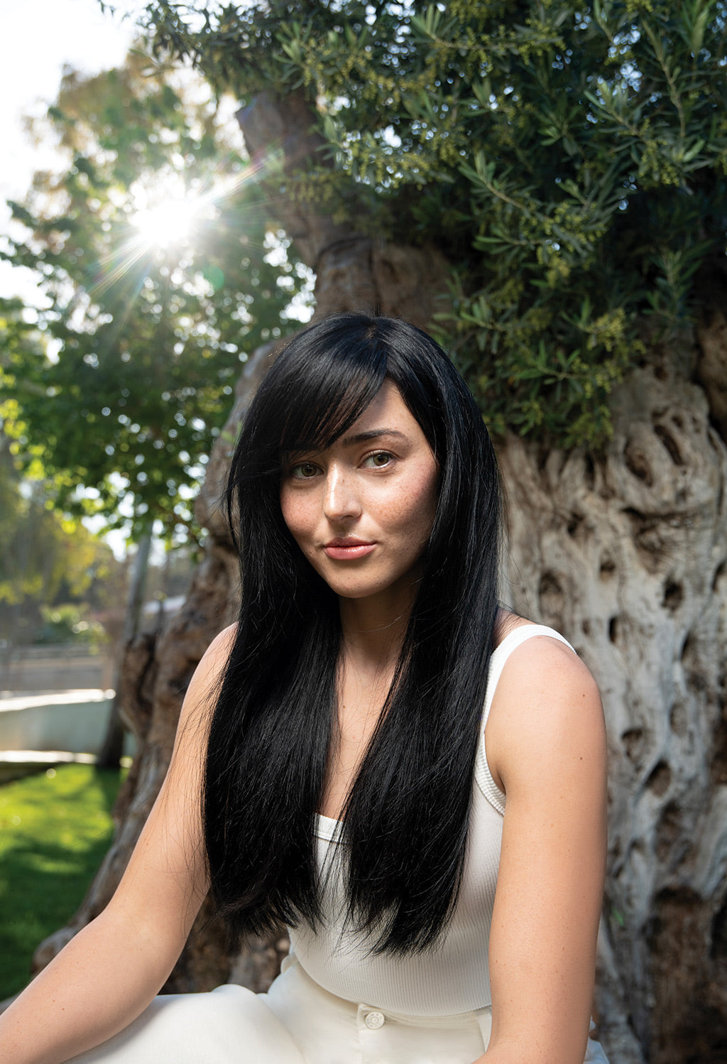 Woman with long black hair sitting outdoors, showcasing smooth and shiny hair in sunlight.