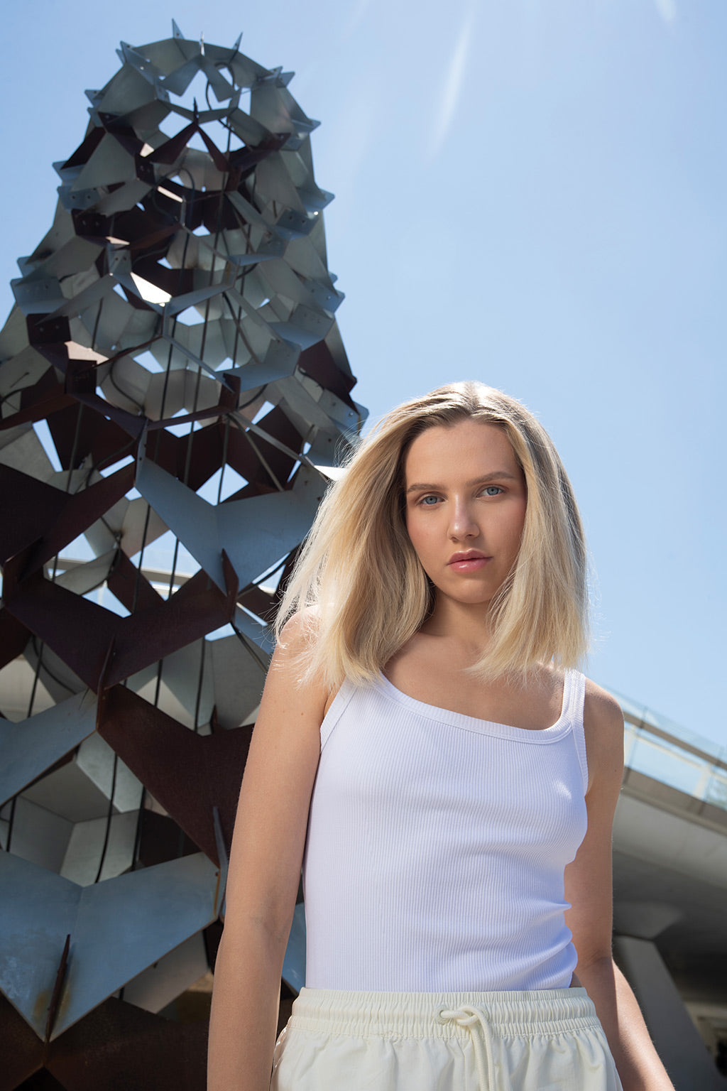 Young woman in a white tank top stands in front of abstract metal sculpture against a clear blue sky.