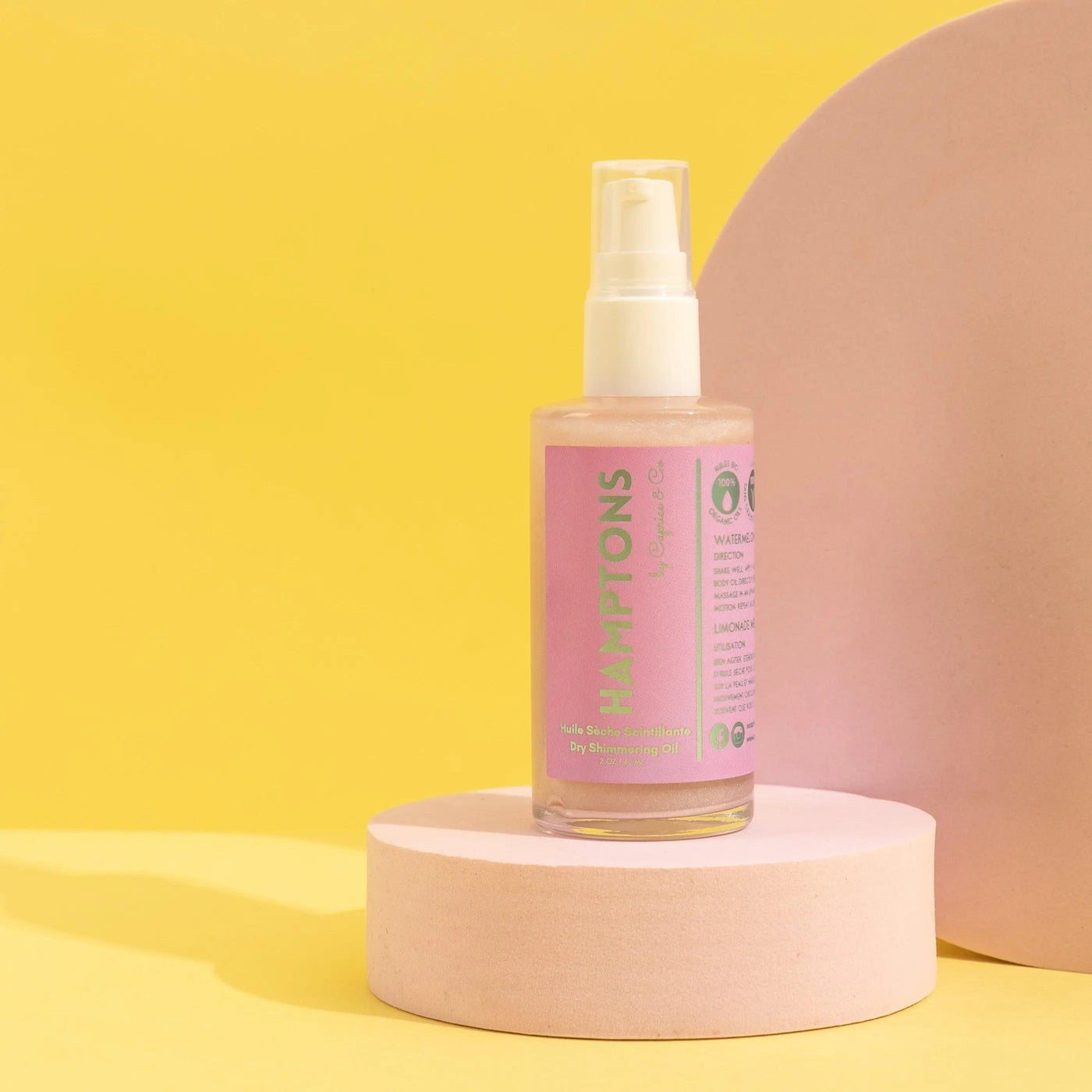 Hamptons Dry Shimmering Oil in a pink bottle on a pastel backdrop, showcasing organic body oil for a glowing finish.