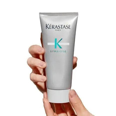 Kérastase Symbiose Scalp Purifying Micro-Peeling Scrub in a silver tube held in a hand, ideal for revitalizing hair health.